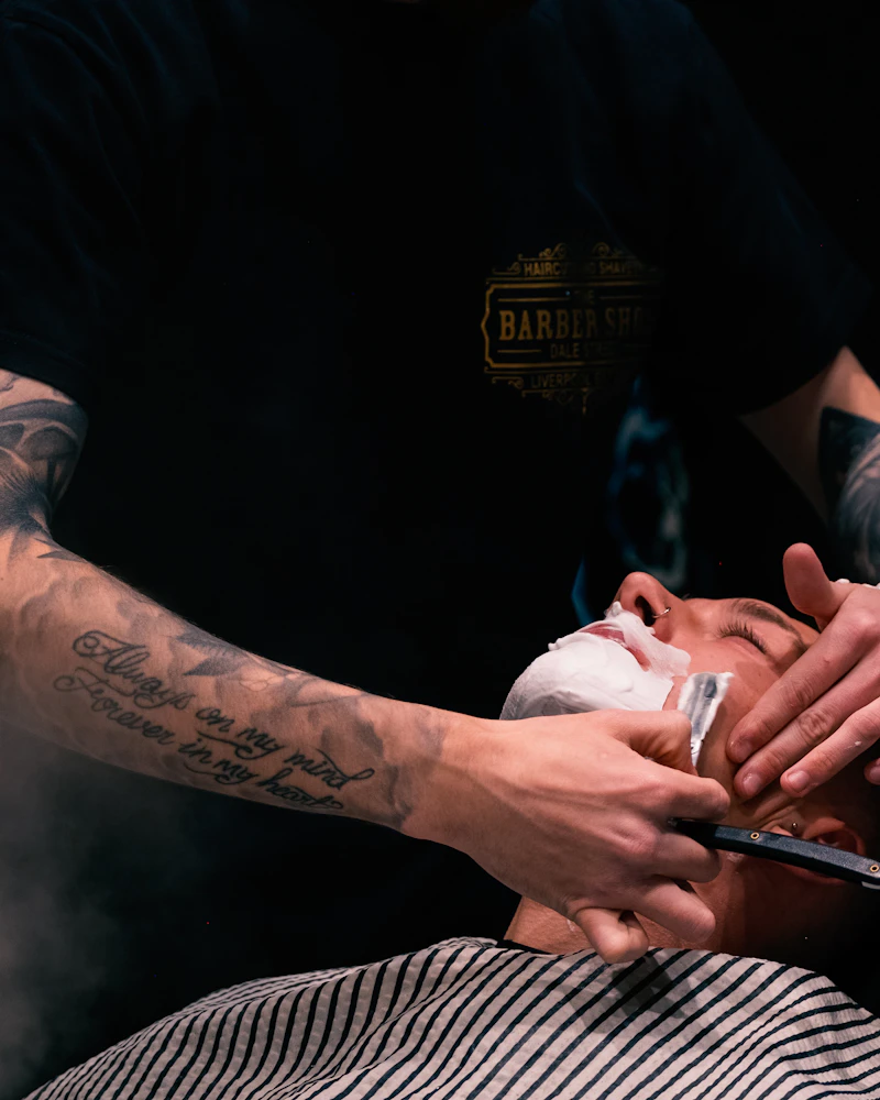 Barber brije mušteriju britvom uz topli ručnik u zamračenom prostoru salona.